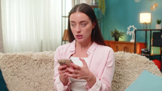 Young woman sitting on home sofa angrily types on smartphone chatting and waves hand in frustration at negative messages. Caucasian girl shows displeased look and posture reacting to words online.