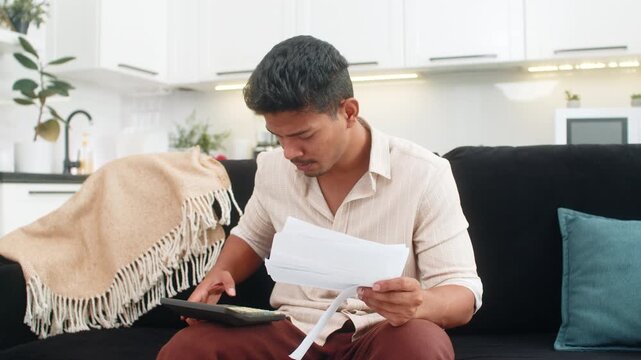 Indian man at home holds utility bills uses calculator enters numbers for budget and squints at totals. Arabian guy on sofa marks priorities writes notes sets payment order plans monthly costs calmly