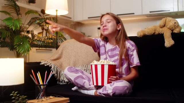 Teenager girl at night home watches film eats popcorn from bowl giggles then bursts into loud laugh. Caucasian child kid on sofa at evening points at screen claps hands enjoys fun story pure big joy.