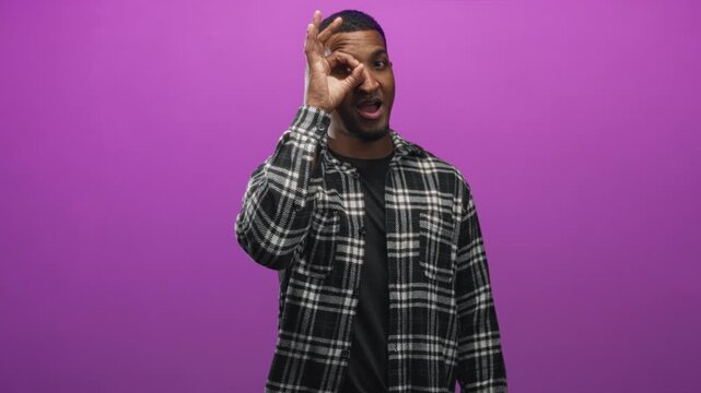 Young black man wearing plaid shirt makes ok sign with hand over eye in studio with purple backdrop; playful curiosity.