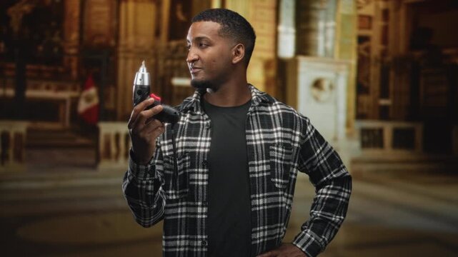 Man holding electric screwdriver with hand on hip inside a building cathedral interior, plaid shirt and focused gaze, peruvian flag visible; contemplation craft.