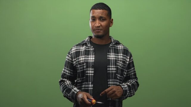 Man holding a screwdriver and pointing it in a studio against a green greenscreen while wearing a plaid flannel shirt, looking toward the tool tip; instructional focus.