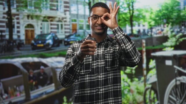 Young black man in plaid shirt and glasses holding takeaway coffee cup while making ok sign to eye on street with canal boats behind; playful curiosity charm.