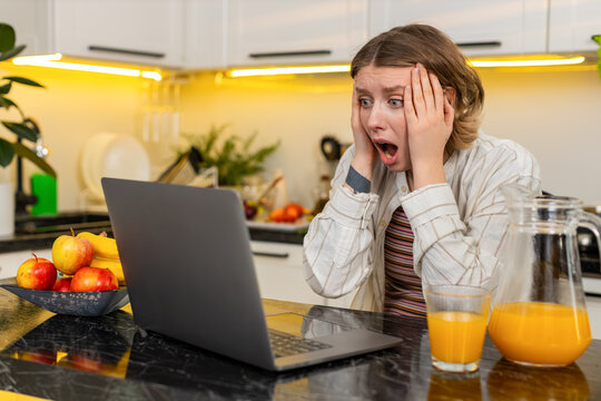 Young woman looks at laptop screen in home kitchen after grim news feeling shock defeat and failure during work. Adult girl struggles with loss stress and disappointment after bad message ruins plans.