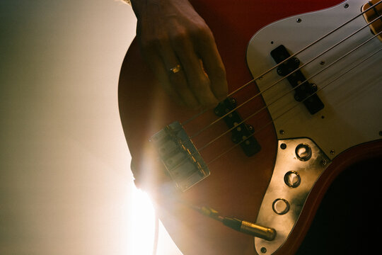 Close-up of a hand plucking strings on an electric bass guitar body with knobs and cable jack, lit by bright backlight.