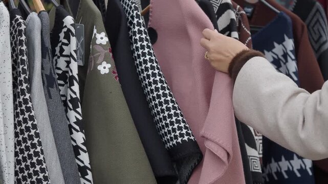 A woman's hands browse through a rack of diverse winter capes and ponchos, examining various colors and patterns for sale in a clothing store.