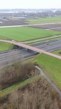Vertical screen Aerial Highway Overpass With Trucks Crossing Green Fields, Slowmoving Cars On Wet Asphalt, Winter Trees Along Drainage Ditch, Drone Pan Showing Transport Corridor And Rural