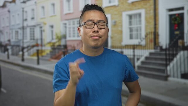 Man in blue shirt and glasses holds up two fingers and points with index finger toward an offframe object on a residential street lined with rowhouses; casual confidence.