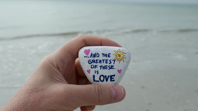 And the greatest of these is love kindness rock in hand with ocean waves in background