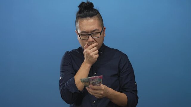 Man counting indonesian rupiah banknotes with visible hands and furrowed brow in blue studio; concern finances planning.