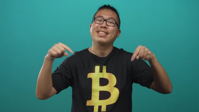 Man pointing fingers to bitcoin t shirt while smiling in a teal studio setting, hands visible and chest logo prominent; cryptocurrency pride.
