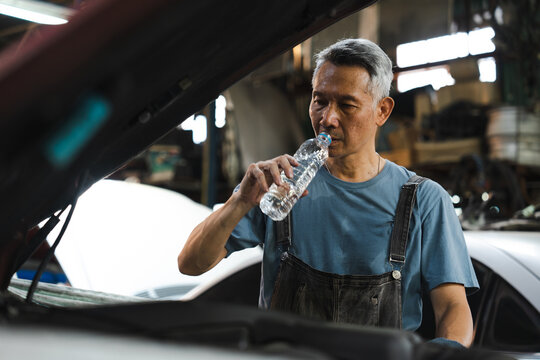 Asian mechanic car repair auto workshop drinking water blue shirt overall senior man break time hydration hot day Calm Asian mechanic blue shirt and overall taking break time for hydration while