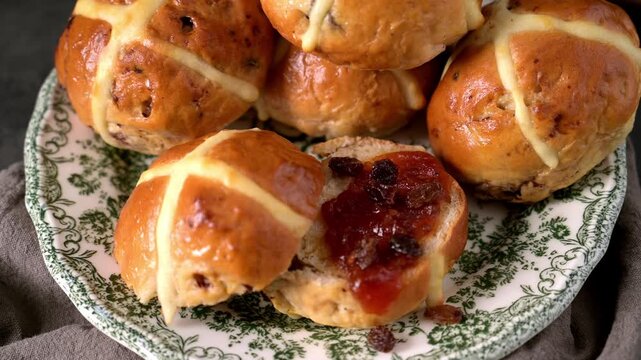 A close up shot of a glazed hot cross bun sliced in half with dark fruit jam and raisins. The buns feature a soft golden texture and are served on a green patterned vintage plate.