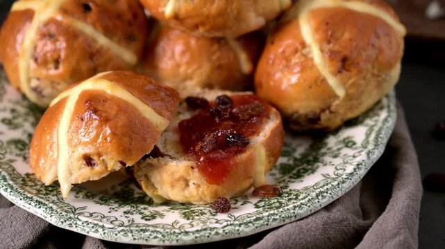 Golden brown glazed hot cross buns served on a patterned ceramic plate. One bun is sliced open to reveal a rich fruit jam filling and scattered raisins under soft natural lighting.