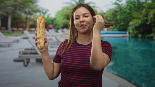 Woman holding baguette while covering face with hand at hotel poolside near lounge chairs and turquoise water; playful moment.