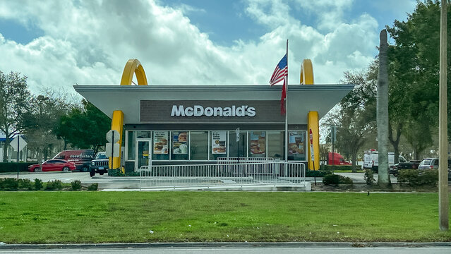McDonald's fast food restaurant exterior with iconic golden arches and American flag in front, located in a suburban area with green grass