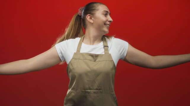 Woman in apron smiling with arms outstretched and clapping motion, wearing white t shirt and ponytail scrunchie against red studio backdrop; joyful welcome.