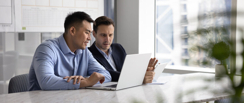 Teamwork on project development. Wide panoramic web banner image two diverse male business coworkers sit together at office table compare data on laptop tablet synchronize information coordinate tasks