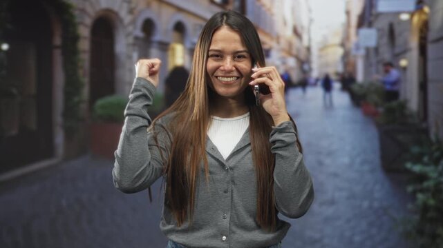 Woman brunette smiling holds smartphone to ear while pumping her fist on a narrow cobblestone street; joy celebration.