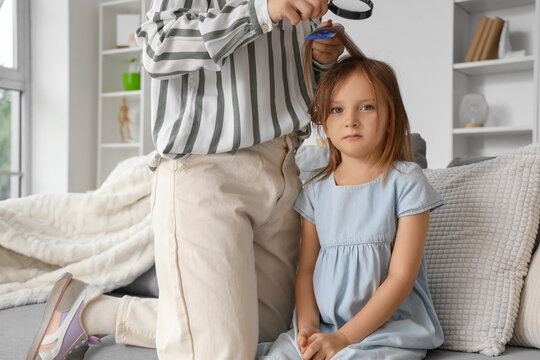Mother checking little girl's hair with magnifying glass and comb for head lice at home, child sitting on sofa during hygiene and health inspection