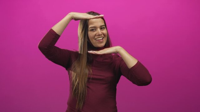 Young hispanic brunette woman framing her face with hands in a studio magenta backdrop, wearing a burgundy turtleneck and smiling at camera; playful confidence.