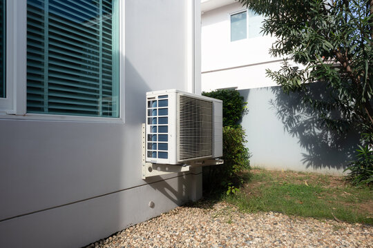 Outdoor air conditioning unit on side of residential house, system mounted on exterior wall, positioned for efficient airflow and effective heat exchange, ensuring comfortable indoor conditions.