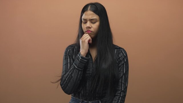 Young latin woman with hand on chin and closed eyes in studio against peach background; concern reflection.