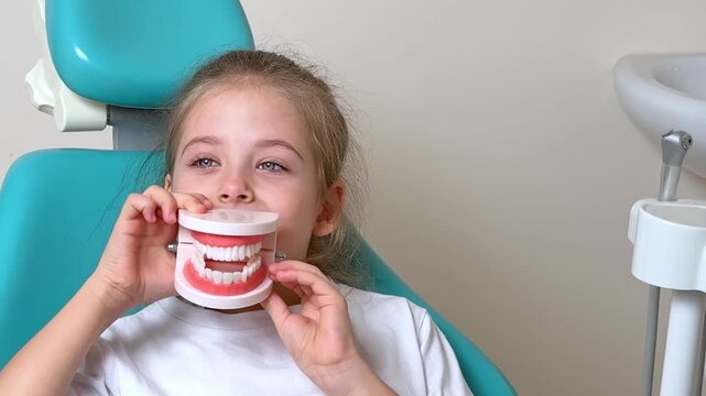 Little girl holding a dental model in front of her mouth while sitting in a dental chair with a light blue background and dental equipment visible. Slow motion footage