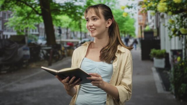 Young woman holding an open book and smiling while reading on a leafy city street, hands visible and casual shirt; quiet study serenity.