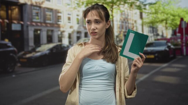 Young woman holding green learner l plate with hand on chest on street, eyes closed; driving anxiety.