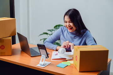 Female entrepreneur packing product box at office with smartphone, laptop representing ecommerce business, online selling, order fulfillment, small business workflow digital marketing startup
