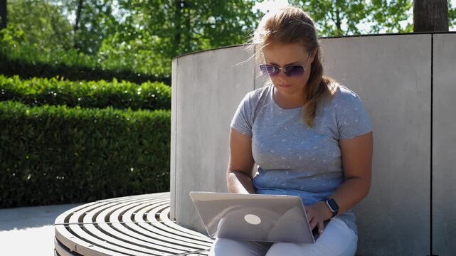 A woman is working on a laptop in a city park. The general layout of a modern park features trimmed bushes, trees, and comfortable benches, on which the woman sits with her laptop.