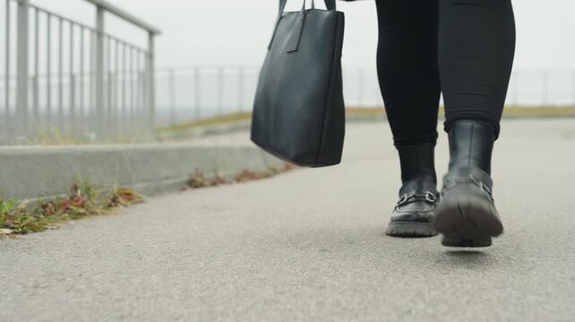 Boots stroll confidently. Walking past railing with style. Steady steps and swinging tote during overcast walk. Confident front view of boots on rhythmic stroll by railing with swinging tote