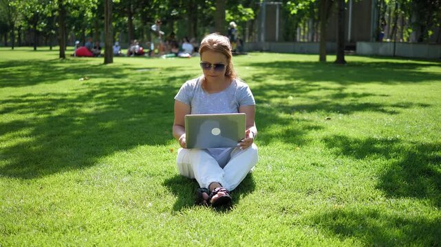 A young woman in sunglasses is working on a laptop in the summer park. She is sitting on the grass. The camera slowly moves around the woman. The video is stabilized.