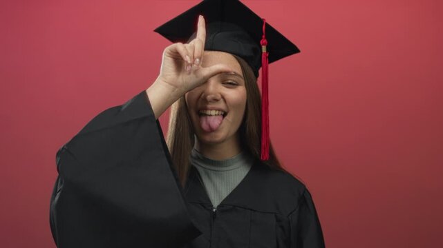 Girl wearing graduation cap sticks tongue out and makes l sign on forehead in studio; playful pride.