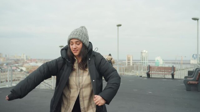 Woman dancing on rooftop wearing beanie, pulling on puffer coat with city skyline backdrop, overcast daylight, playful solo movement and candid joyful expression, improvisational street dancer vibe