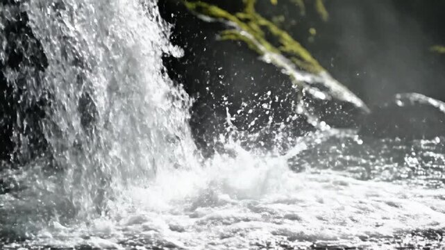 Close-up of water splashing over rocks in a natural waterfall, with green moss and foliage visible in the background, evoking a sense of fresh, wild nature