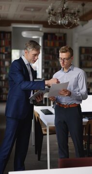 Two businessmen, office employees working on contract terms, discussing project data holding documents and digital tablet, shaking hands, finalizing collaboration meeting in workplace. Business deal