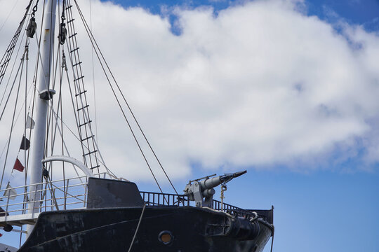 Historic black ship bow with prominent mast, rigging and a deck gun against a blue sky and clouds