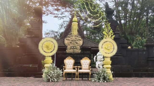 Elegant Balinese Janur Wedding Gate with Morning Sunlight, Traditional Palm Leaf Decorations, Golden Chairs, Floral Arrangement and Cultural Archway in Tropical Outdoor Ceremony Setting