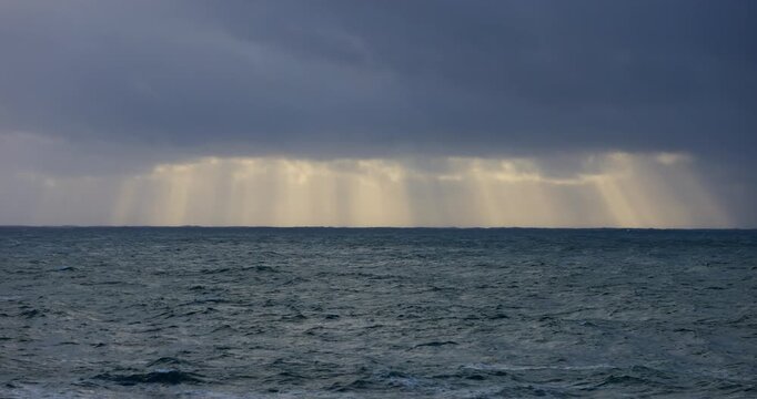 A rough stormy ocean, with moody clouds and bright sun rays shining down on the horizon. Captured in slow motion from a coastal headland.