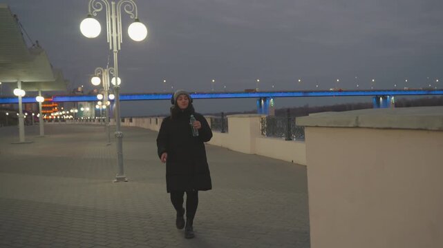 Dusk riverfront solo dance by lampposts. Woman in dark parka and beanie strolls along cobblestone promenade, holding bottle, pausing at railing bluelit bridge and distant city lights create calm,