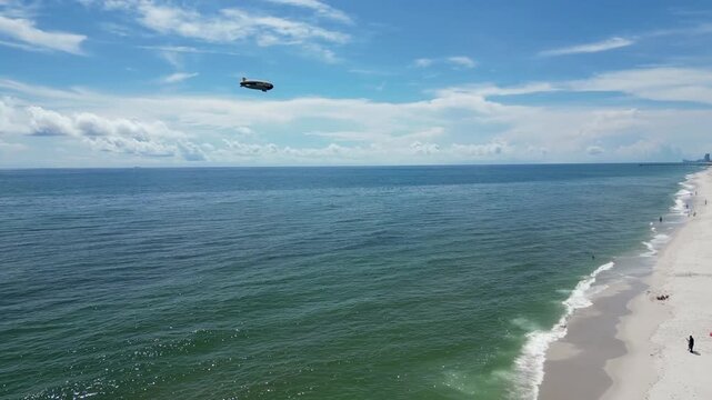 Drone footage of a blimp in a blue sky over the sea shore. People relax on the sand as waves hit the coast. Great for travel and tourism commercials.