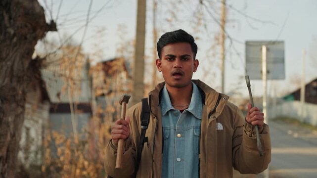 Closeup man holding hammer and backpack on roadside, intense gaze and poised hands, cracked pavement and street sign behind, autumn foliage and low evening light, confident tradesman ready to fix