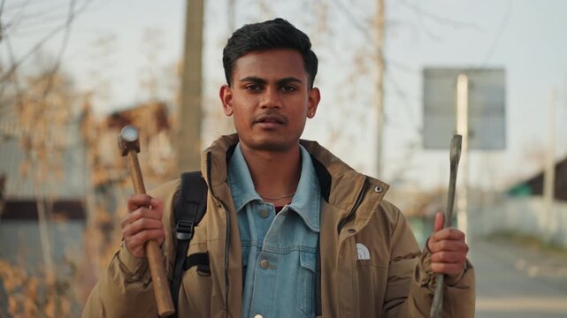 Young man holding hammer and chisel outdoors, closeup portrait with firm grip, denim shirt under jacket, backpack visible, autumn roadside backdrop, instructional demonstration vibe, confident