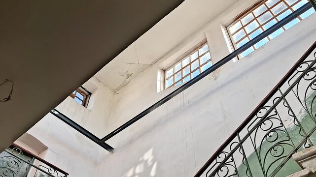 Upward view of stairwell with ornate metal railings, stone steps, and natural light through large windows, emphasizing architectural details, shadows, and structural features