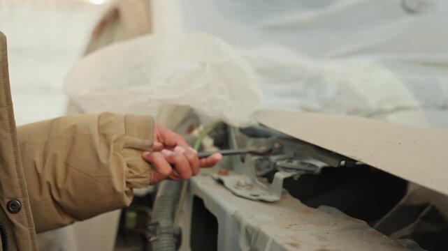 Closeup damaged hood with plastic cover, exposed wiring and bent frame, inspector role assessing clips and cables, dirt and dust, careful documentation and estimate prep, muted outdoor salvage