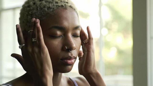 A diverse person in a muted camisole gently massages their face with naturally glowing skin, slowly opening eyes to a camera with a soft smile, bathed in soft high-key light against a creamy bokeh