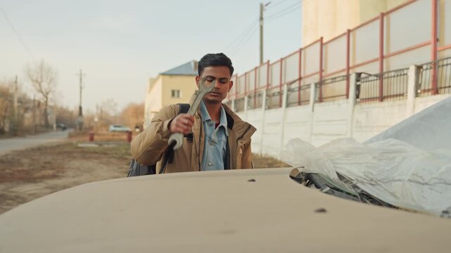 Young man inspecting damaged car hood outdoors, backpack slung, suburban dirt road, missing front bumper, exposed engine bay, concerned expression, hands on edge of hood, muted autumn light, preparing