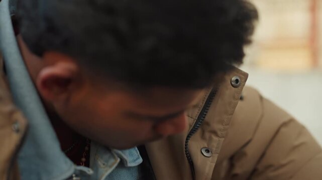 Young man inspecting engine with wrench, leaning over damaged hood in outdoor scrapyard closeup of hands tightening bolt, concentrated expression, denim shirt under brown jacket, methodical